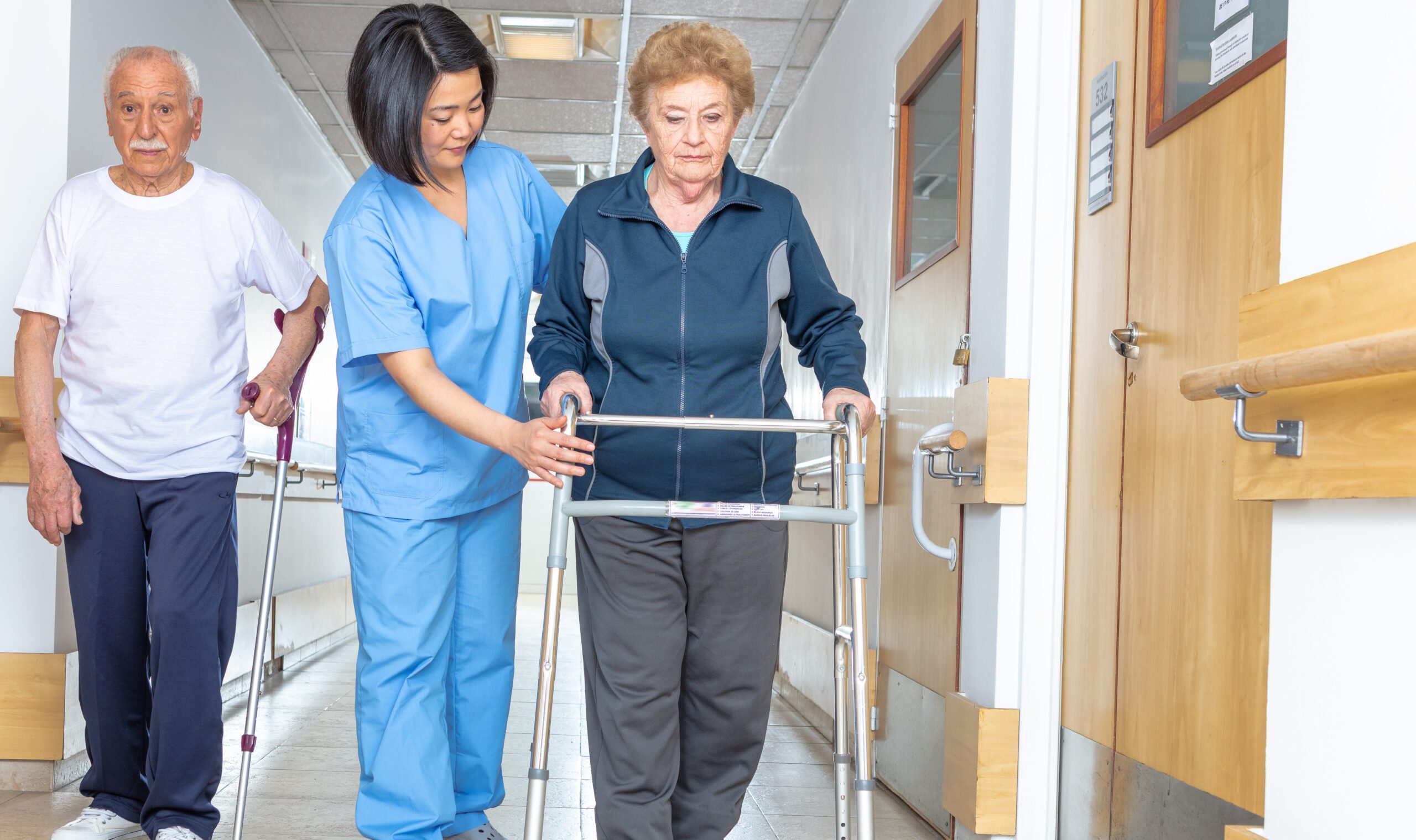 Asian doctor helping elder woman with walker and man in hospital aisle